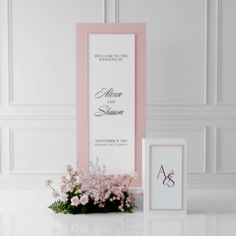 Framed wedding welcome sign with pink border and floral arrangement on a white wall.