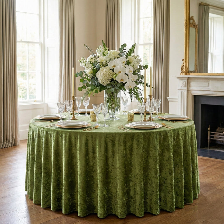 Elegant dining table setting with a sage green velvet tablecloth, white flowers, and glassware in a room with large windows and a fireplace.