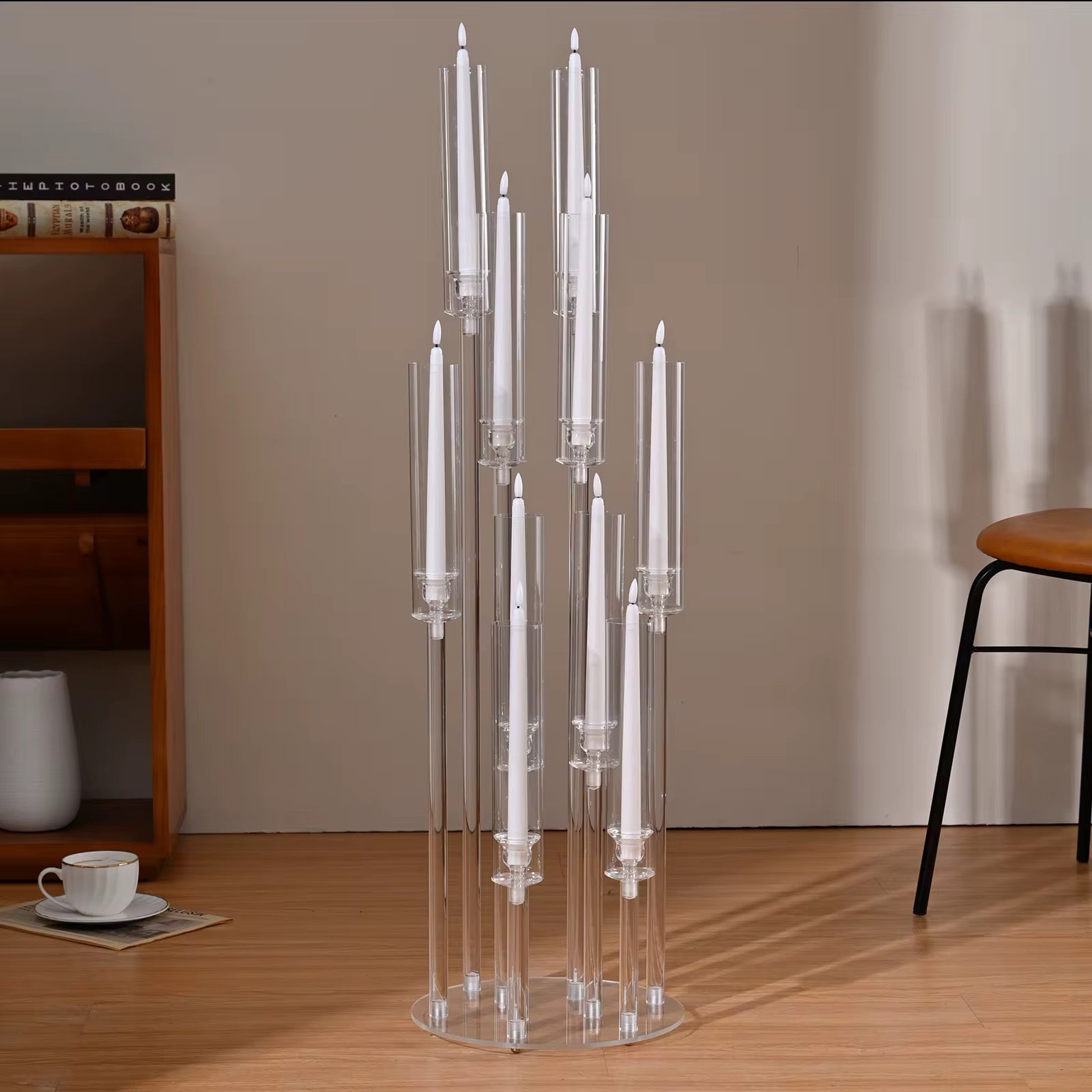 Clear candle holder with white candles on a wooden floor, with a chair and shelf in the background.