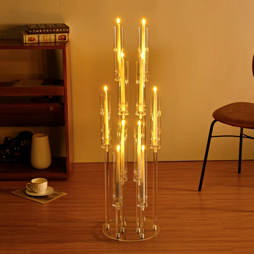 Clear glass candle holder with multiple candles on a wooden floor, with a bookshelf and chair in the background.