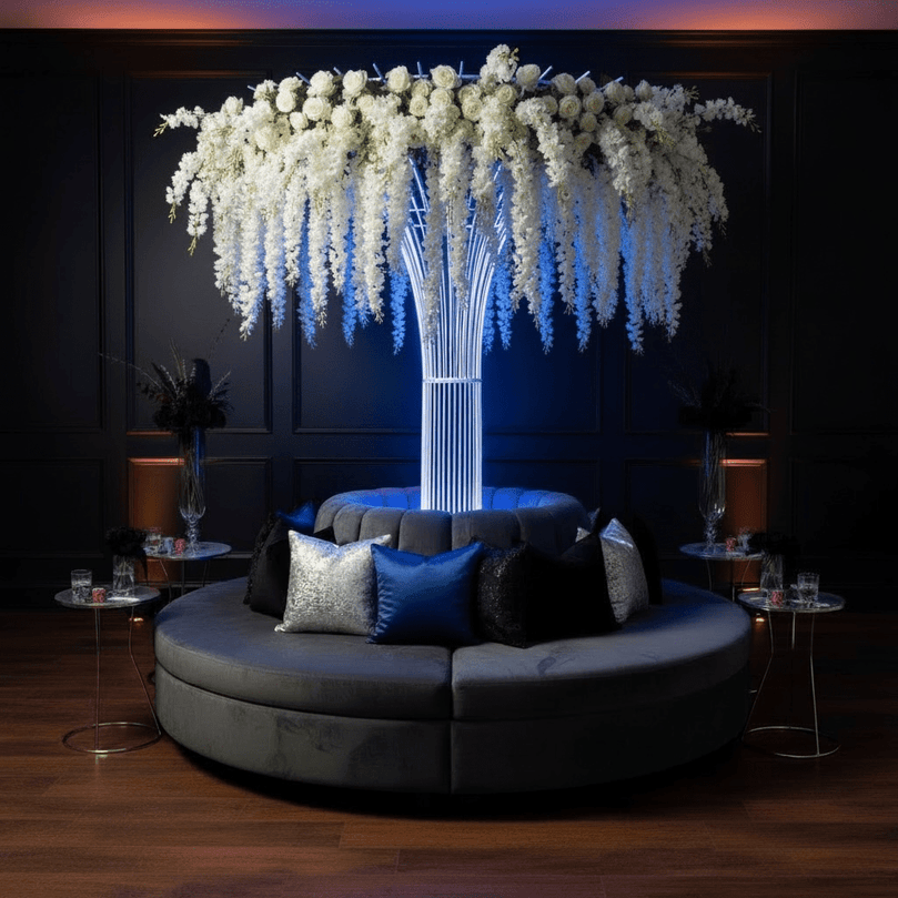 Modern circular sofa with decorative pillows in a dimly lit room with a large floral arrangement tied to a white wedding rental prop stand.