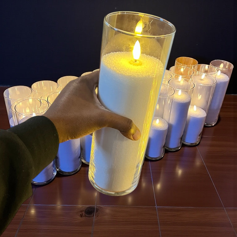 Hand holding a tall, lit candle in a glass container with more candles in the background on a wooden surface.