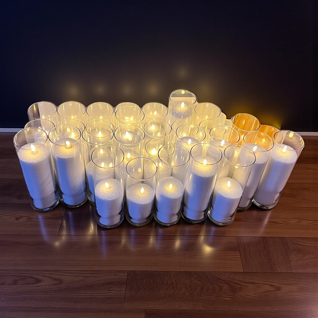 Multiple white candles in glass holders arranged on a wooden surface with a dark background.