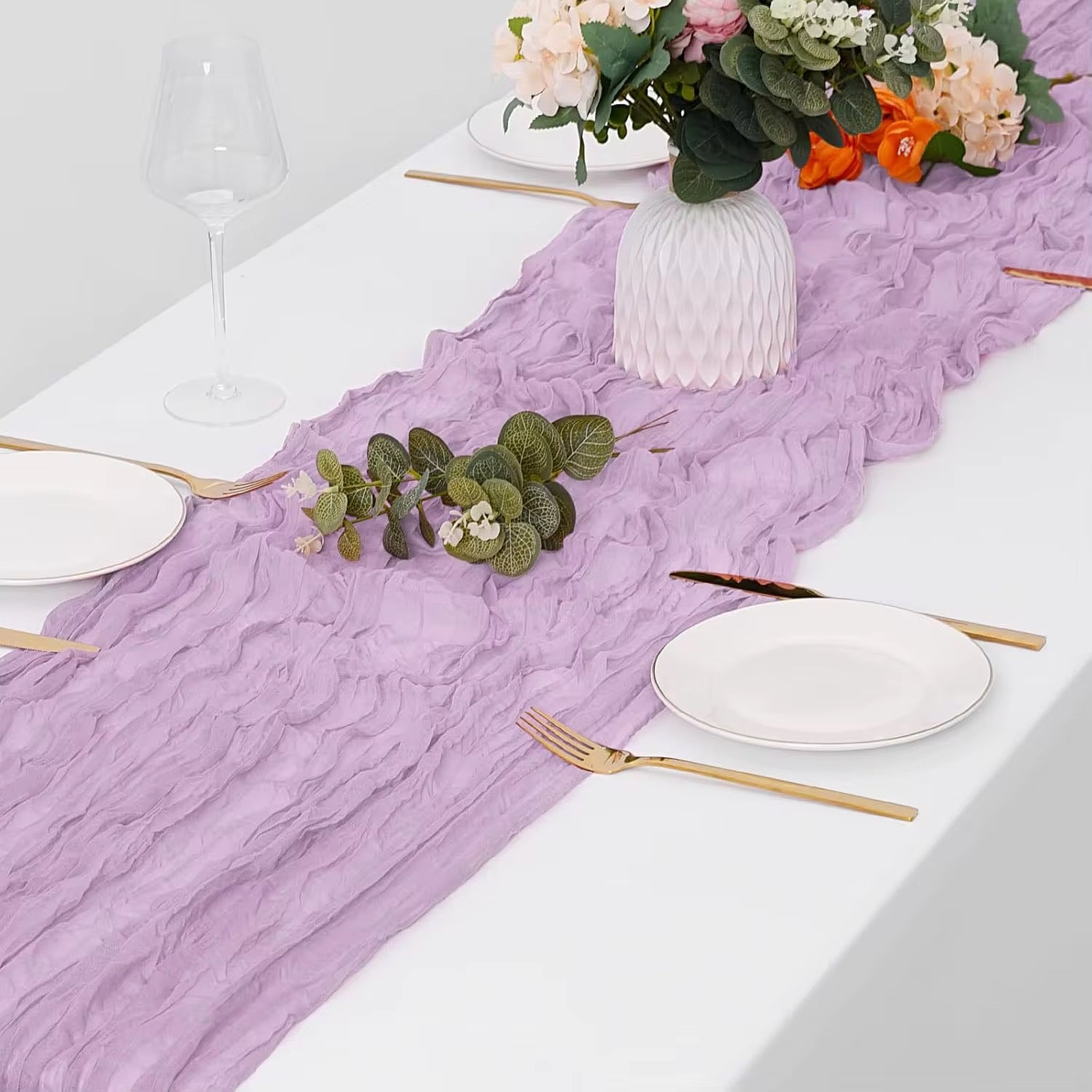 Table setting with a light purple cheesecloth table runner, white plates, gold cutlery, and floral decorations.