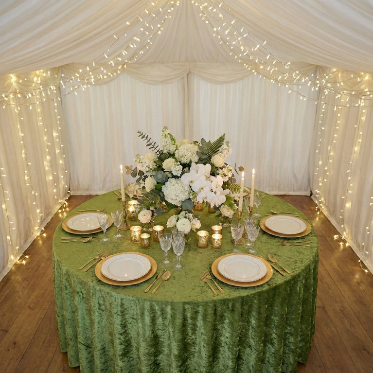Decorated table with sage green velvet tablecloth, flowers and candles inside a tent with string lights