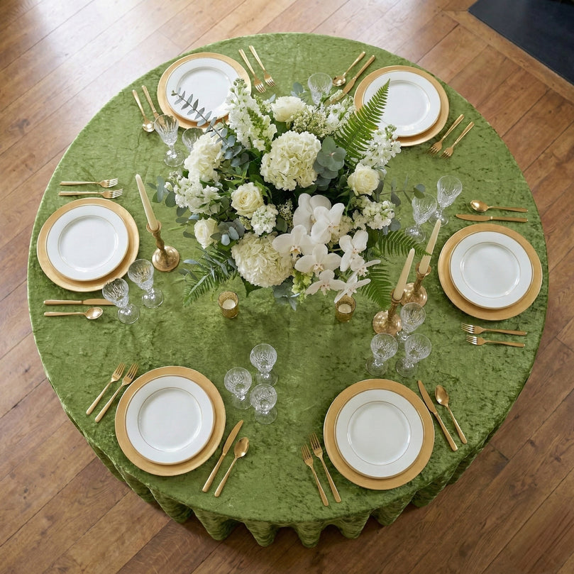 Elegant table setting with white plates, gold chargers, and sage green velvet tablecloth on a wooden floor.