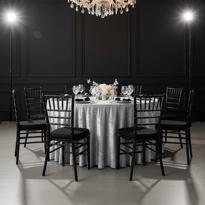 Dining table set with silver velvet tablecloth and black chiavari chairs in a dimly lit room.